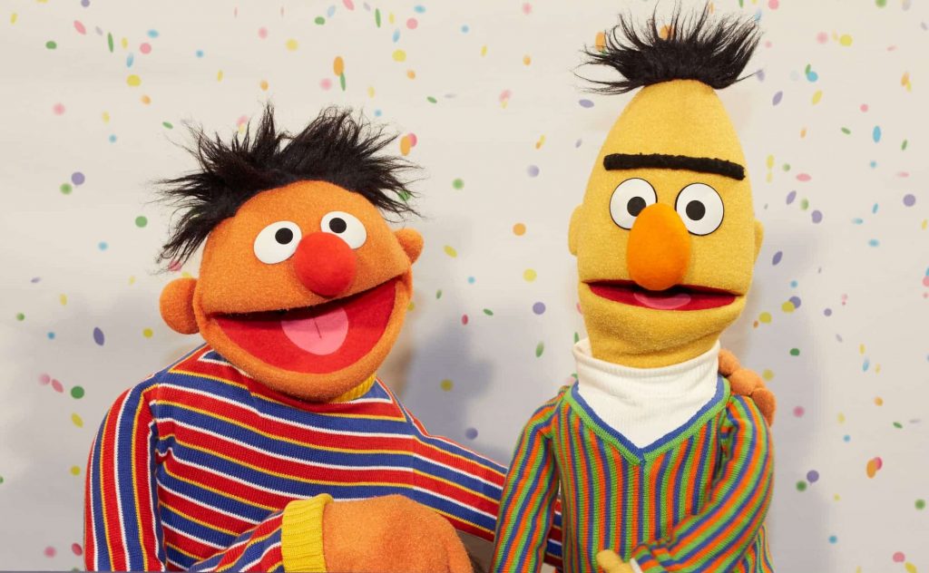 Bert and Ernie from Sesame Street. The orange and yellow muppets are wearing brightly striped clothing and are holding each other.
